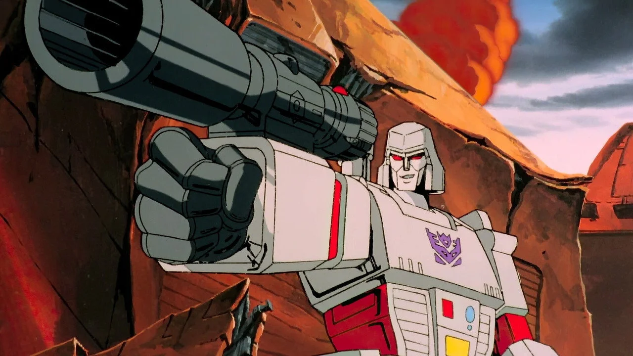Megatron on a red rock ridge with his arm cannon extended toward the camera, purple Decepticon badge on his chest, smoke in the sky behind