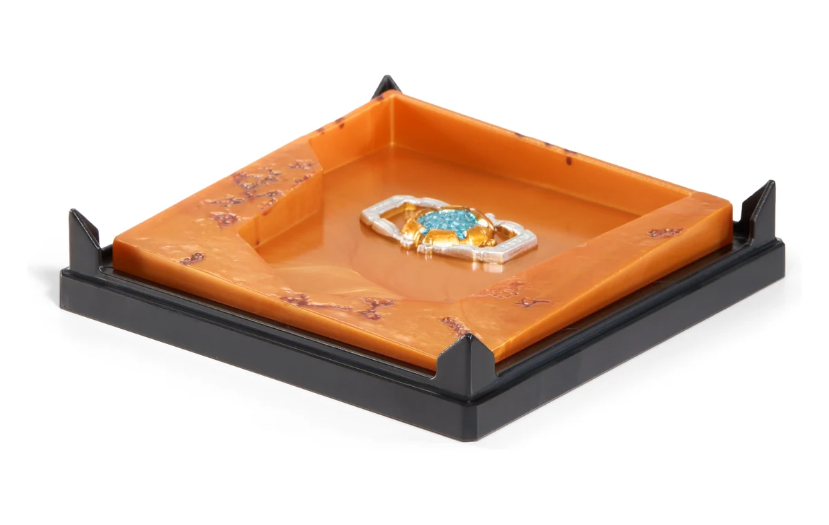 The diorama base photographed empty, a square of orange plating in a black frame with a recess where the figure sits. Set into the recess is a sculpted Matrix of Leadership in silver and gold with a blue core.