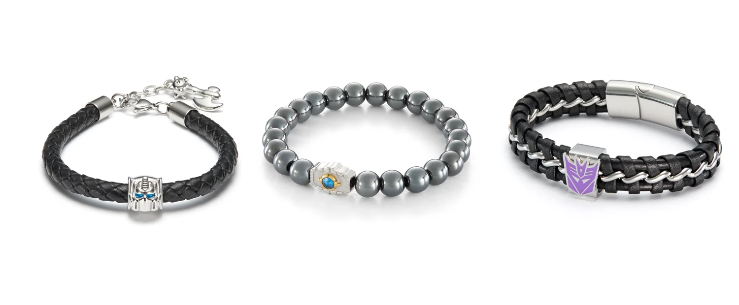 Three Transformers bracelets on white. Left, a braided black leather band with a silver Optimus Prime face bead and a small silver Energon axe hanging from the extension chain. Centre, a strand of dark grey hematite beads with a silver and gold Matrix of Leadership charm holding a blue stone. Right, a braided black leatherette band with a silver chain running through it, a purple Decepticon shield charm and a silver slide lock closure.