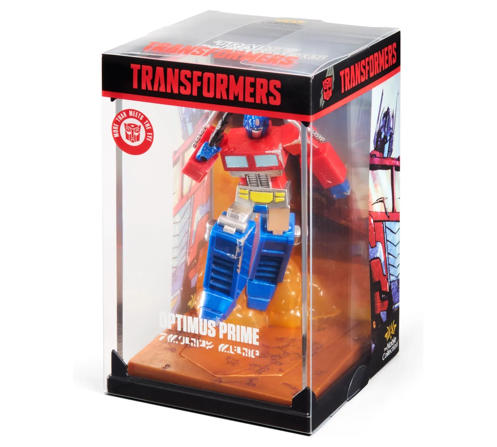 The Optimus Prime diorama in its retail package, a clear acrylic case over a black base. A red band across the top carries TRANSFORMERS in white, a red roundel on the left reads MORE THAN MEETS THE EYE around an Autobot sigil, a printed city backdrop sits behind the figure, and the Noble Collection logo is in the bottom corner.