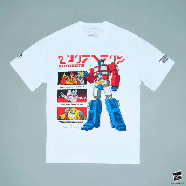A white oversized t-shirt laid flat. G1 Optimus Prime stands the full height of the print in red, blue and grey, with red Japanese katakana and the word AUTOBOTS above him. Three stacked panels run down the left of the print showing Hot Rod, Arcee and Bumblebee, captioned EVERY HERO HAS A BEGINNING and MORE THAN MEETS THE EYE, with TRANSFORMERS x WOLFPAK in pale grey beneath them.
