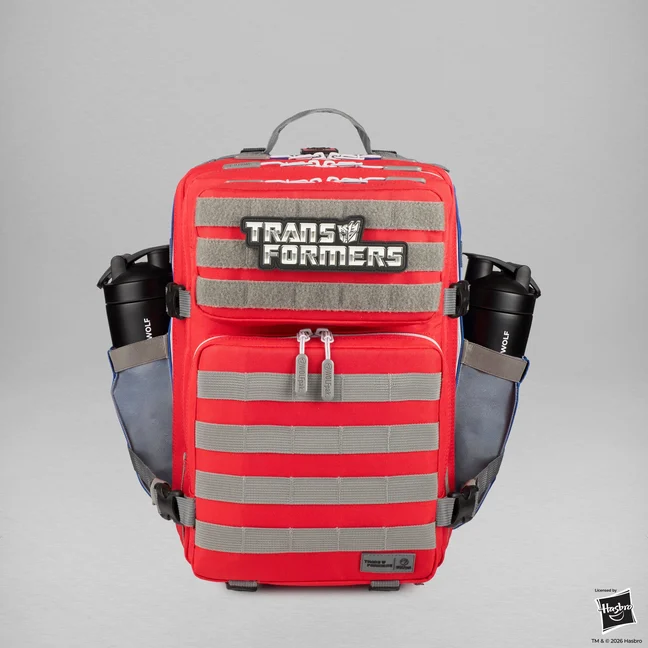 A red tactical backpack photographed square on against grey. Grey hook and loop panels and grey MOLLE webbing run across the red fabric in horizontal bands, a raised black and silver TRANSFORMERS patch sits on the upper panel, blue trim shows along the edges, and a black WOLFpak shaker bottle sits in each side pocket.