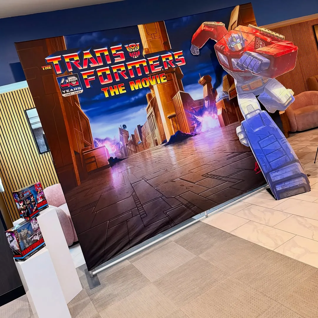 Lobby backdrop for The Transformers The Movie 40th anniversary, a painted Optimus Prime reaching out of a Cybertronian street scene, with boxed figures displayed on white plinths in front of it