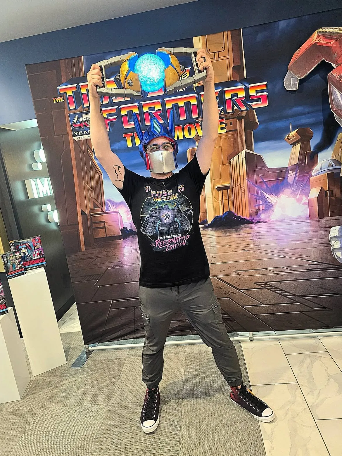 A fan wearing an Optimus Prime helmet with a silver faceplate and a Reformatted Edition soundtrack t-shirt, holding a lit Matrix of Leadership replica above his head in front of the 40th anniversary backdrop
