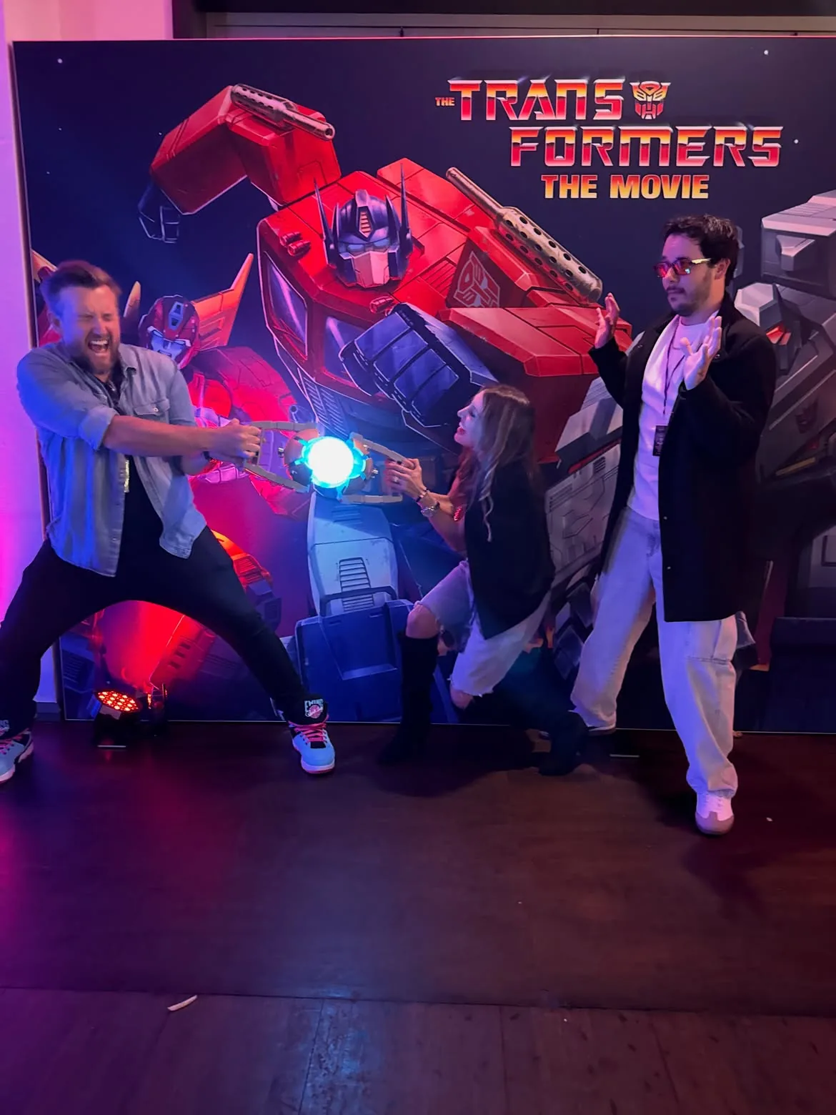 Three guests staging a tug of war over a lit Matrix of Leadership replica in front of a large Optimus Prime and Megatron standee, one pulling hard with his mouth open, one kneeling and grabbing the middle, one standing back with his hands up