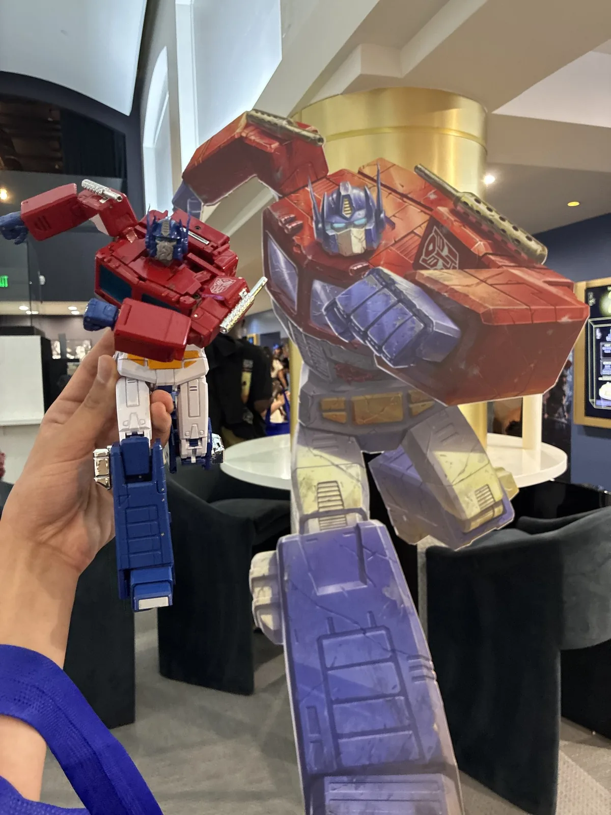 A hand holding an Optimus Prime action figure up in the air so that it lines up with a much larger Optimus Prime standee behind it