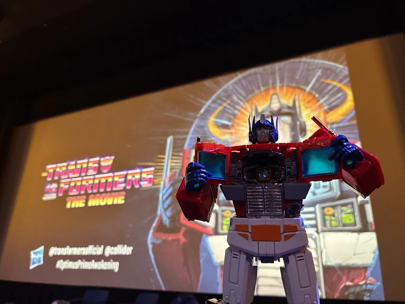 A cinema screen showing an Optimus Prime figure against a glowing orange background with The Transformers The Movie logo and the handles at transformersofficial, at collider and the hashtag OptimusPrimeAwakening