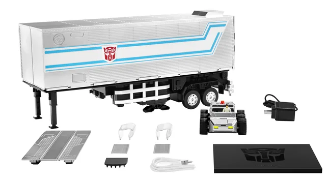 The Robosen Optimus Prime Trailer Kit laid out on white: the silver and white trailer with blue stripes and a red Autobot sigil on its flank, two metal ramp plates, two white support cradles, a grey pad, a small black connector block, a white USB-C cable, a black power adapter, the Roller scout car with a Spike figure beside it, and a black display base with the Autobot sigil embossed into it.