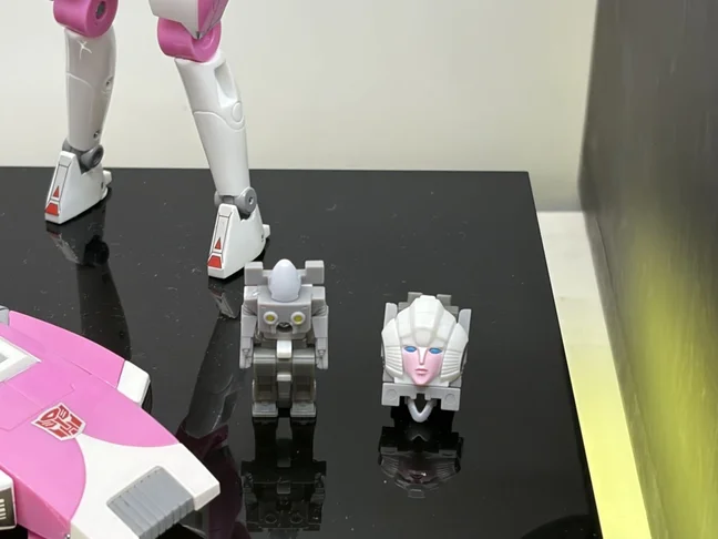 Two small grey units stand on the black display plinth beside Arcee's lower legs and the nose of her pink car: a tiny robot with yellow eyes on the left, and her own white face folded into head mode on the right