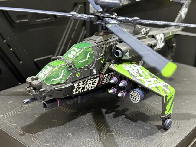 The Overgear OG-02 EX Grinder colour sample in helicopter mode on a dark plinth: a black gunship with an acid-green translucent canopy, green stub wings carrying white brush-stroke kanji, pink-tipped missiles and a rocket pod