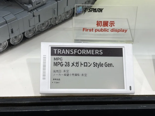 A Takara Tomy display card reading TRANSFORMERS, MPG, MPG-28 Megatron Style Gen., with release date and retail price both listed as undecided. The tread of a grey tank sits behind it, under an acrylic plaque reading First public display