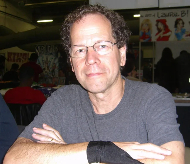 Bob Budiansky seated with his arms folded behind a table on a comic convention floor, wearing glasses and a grey t-shirt, with other exhibitors and artwork behind him.