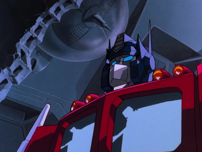 Optimus Prime mid-transformation, his blue helmet and silver faceplate risen above the red cab of his truck mode, orange marker lights along the roof, grey machinery behind him.