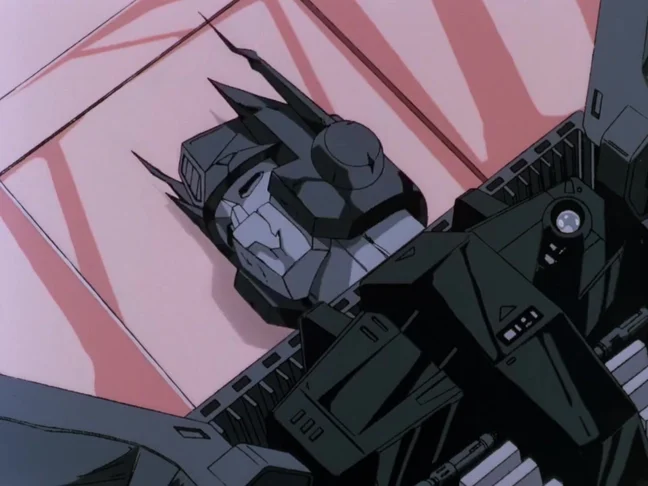 Optimus Prime drained of all colour to a flat grey, head tilted back and body slumped against a pale pink wall in the Autobot medical bay.