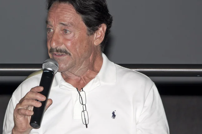 Peter Cullen standing at a microphone on a darkened stage at the Kennedy Space Center in 2011, wearing a white polo shirt.