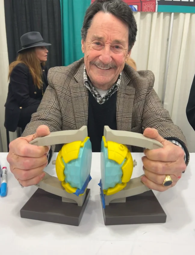 Peter Cullen at a convention table in 2024, smiling, holding up an open replica Matrix of Leadership with both hands.