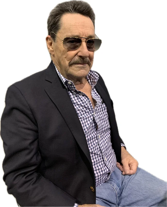 Peter Cullen photographed seated in a dark blazer over a purple and white checked shirt and jeans, wearing gold aviator sunglasses, cut out against a white background