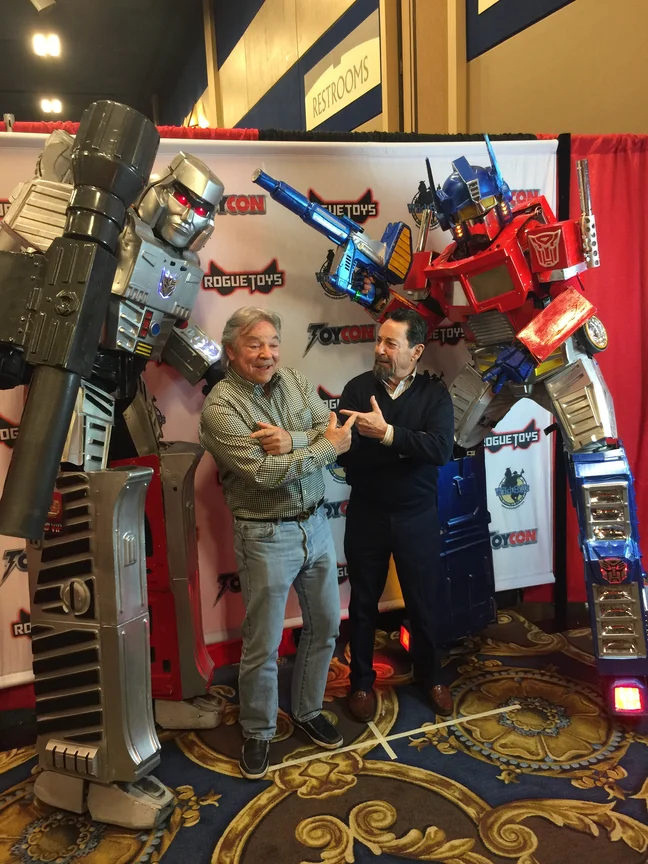Frank Welker and Peter Cullen pointing at each other at a RogueToys ToyCon, standing in front of life size Megatron and Optimus Prime statues